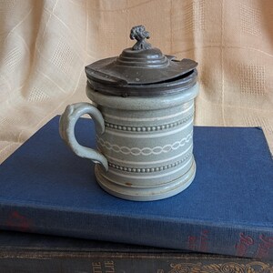 May include: A vintage ceramic salt cellar with a pewter lid. The cellar is decorated with a white and gray pattern and has a handle.