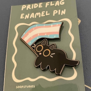 Puede incluir: Un pin de esmalte negro con un gato sosteniendo una bandera del orgullo transgénero. El pin está en una tarjeta verde que dice "PRIDE FLAG ENAMEL PIN" y "SOOPSTUDIOS". La bandera tiene rayas celeste, rosa y blancas.
