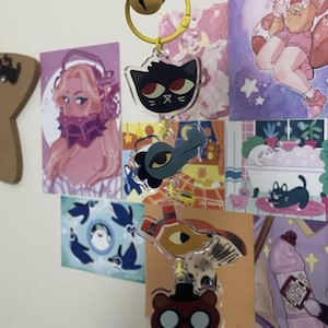 Könnte beinhalten: Eine Sammlung von Acryl-Schlüsselanhängern mit Cartoon-Tierfiguren. Die Schlüsselanhänger sind an einer gelben Metallglocke befestigt. Zu den Figuren gehören eine schwarze Katze, eine blaue Kreatur, eine vogelartige Figur und ein Bär mit Brille.