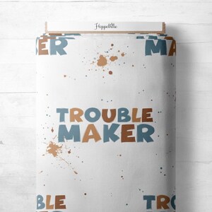 Könnte beinhalten: Eine Rolle weißen Stoffes mit dem Aufdruck "TROUBLE MAKER" in Braun und Blau. Braune Farbspritzer und das Wort "Hoppelhase" sind ebenfalls zu sehen. Geeignet für Bastel- oder Nähprojekte.