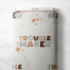 Könnte beinhalten: Gerollter Stoff mit den Worten "TROUBLE MAKER" in braunen und grauen Buchstaben. Braune Farbspritzer sind zu sehen. Die Wörter "ER" und "MA" sind teilweise sichtbar. Der Markenname "Hoppelhase" ist oben aufgedruckt.