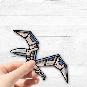 Könnte beinhalten: Ein beige-blauer Papierschnitt eines stilisierten Pterodactylus mit mechanischem Design. Der Ausschnitt wird in einer Hand gehalten.