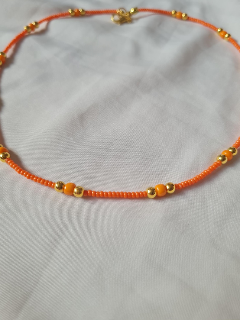 May include: A delicate orange and gold beaded necklace. The necklace is made with small orange beads and larger gold beads.