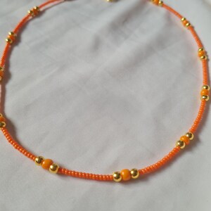 May include: A delicate orange and gold beaded necklace. The necklace is made with small orange beads and larger gold beads.