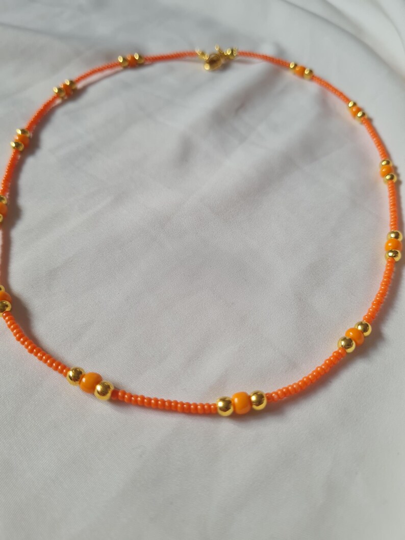 May include: A delicate orange and gold beaded necklace. The necklace is made with small orange beads and larger gold beads.