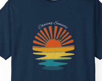 May include: Navy blue t-shirt featuring a sunset graphic with the text "Chasing Sunsets". The graphic includes a sun with orange and yellow rays, reflected in the water below. The shirt is a classic crew neck style.