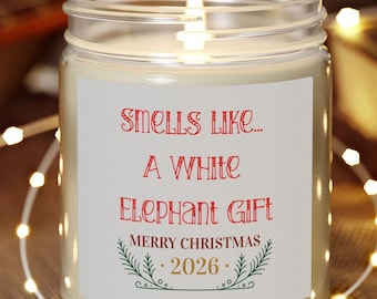 Könnte beinhalten: Eine Kerze im klaren Glasgefäß mit einem weißen Etikett, das in roter und grüner Schrift den Text trägt: Smells Like... A White Elephant Gift Merry Christmas 2026. Die Kerze steht auf einem runden Holzsockel und hat einen brennenden Docht.
