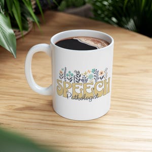 May include: White ceramic mug featuring the text "SPEECH Pathologist" in a retro style, decorated with floral designs in blue, orange, and green. The mug contains dark coffee and is placed on a wooden surface.