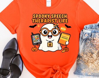 May include: Bright orange t-shirt with a Halloween design. The graphic shows a ghost character wearing glasses and holding a phone and a book. The text "Spooky Speech Therapist Life" is printed above the ghost.