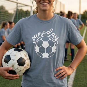 May include: A slate blue short-sleeved t-shirt featuring a white graphic of a soccer ball and the script text "gameday" printed on the front. The shirt has a crew neckline and a standard fit, showcasing a clean design for sports enthusiasts.