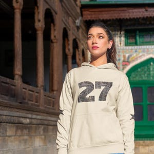 May include: A woman wearing a light beige hoodie with a black and white camouflage print of the number "27" on the front. She is wearing blue jeans and has her hair pulled back.