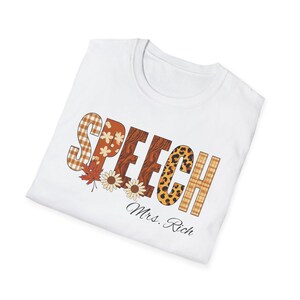Può includere: T-shirt bianca con la scritta "SPEECH" in un design a tema autunnale. Le lettere presentano motivi a quadri, floreali e leopardati. La maglietta include anche il testo "Mrs. Rich" in corsivo.