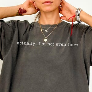Model is wearing a pepper Comfort Colors unisex t-shirt with minimalist centered text reading "actually, I'm not even here".