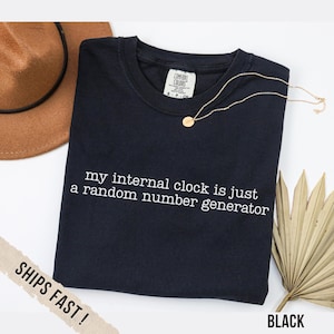 Puede incluir: Camiseta negra con el texto blanco "my internal clock is just a random number generator". Un collar dorado y un sombrero marrón también están en la imagen. La camiseta está etiquetada como "Comfort Colors".