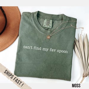 May include: A moss green t-shirt with the text "can't find my fav spoon" in white. A gold necklace with a pendant rests on the shirt. The shirt is folded and placed on a white surface.