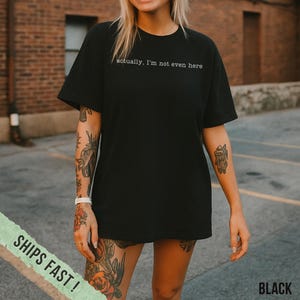 May include: Black t-shirt with the text "actually, I'm not even here" in a simple, white font. The shirt is a basic crew neck style. The word "BLACK" is printed in white on the bottom right corner of the image.