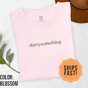 May include: A light pink t-shirt with the word "thirtysomething" printed in black. The shirt is labeled "Comfort Colors" and is a size L-G. The text "COLOR: BLOSSOM" is visible. An orange sticker with "SHIPS FAST!" is also present.