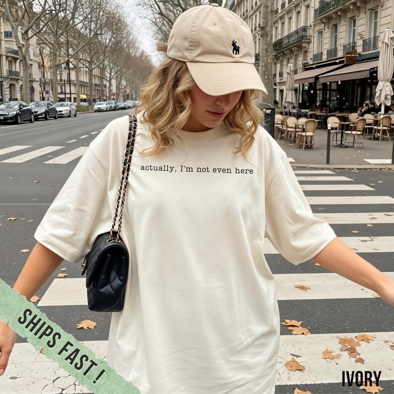 May include: Ivory-colored t-shirt with the text "actually, I'm not even here" in black. The shirt is paired with a beige baseball cap featuring a black logo. A black quilted shoulder bag with a chain strap completes the look.