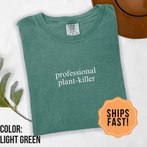 May include: Light green t-shirt with the text "professional plant-killer" in white. The shirt is folded and has a round neckline. A brown hat is visible in the background. An orange sticker with the text "SHIPS FAST!" is in the bottom right corner.