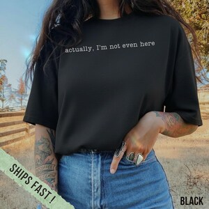 May include: Black t-shirt with the text "actually, I'm not even here" in white. The shirt is a crew neck style and is paired with blue jeans. The image is taken outdoors.