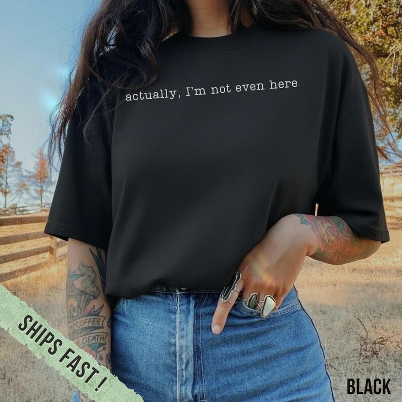 May include: Black t-shirt with the text "actually, I'm not even here" in white. The shirt is a crew neck style and is paired with blue jeans. The image is taken outdoors.