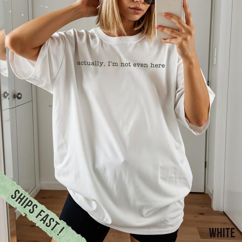 May include: White oversized t-shirt with the text "actually, I'm not even here" in black. The shirt is paired with black shorts. The image also includes a green banner that says "SHIPS FAST!" and the word "WHITE".