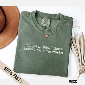 May include: A moss green t-shirt with the white text "sorry I'm late, I don't know how time works". The shirt has a crew neck and short sleeves. A gold necklace with a pendant is on top of the shirt.