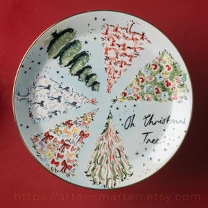 May include: A round, light blue decorative plate with a gold rim. The plate features six stylized Christmas trees in various designs and colors. The words "Oh Christmas Tree" are written in black script. The plate is decorated with small black stars.