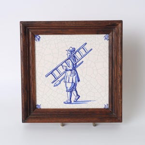 May include: A framed tile featuring a blue and white illustration of a person carrying a ladder. The tile has a crackle effect and is set in a dark wood frame. Decorative corner details are visible.
