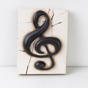 Könnte beinhalten: Ein rechteckiges Wandkunstwerk mit einem schwarzen Violinschlüssel vor einem cremefarbenen Hintergrund mit einer rissigen Textur. Das Musiksymbol ist erhaben und erzeugt einen 3D-Effekt. Das Kunstwerk ist ein dekorativer Gegenstand.