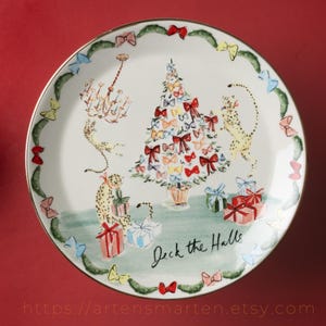 May include: A white ceramic plate with a gold rim, featuring a Christmas tree decorated with bows, cheetahs, and wrapped gifts. The plate is adorned with a garland of bows and the text "Deck the Halls".