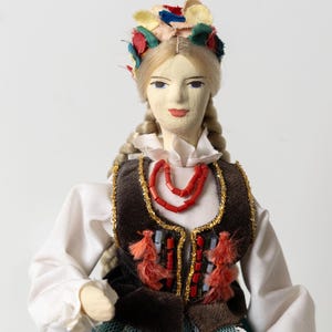 May include: A Polish folk doll with blonde plaits and a colourful floral headdress. The doll wears a white blouse, a brown vest with gold trim, and a red beaded necklace. The vest is adorned with red tassels and decorative elements.