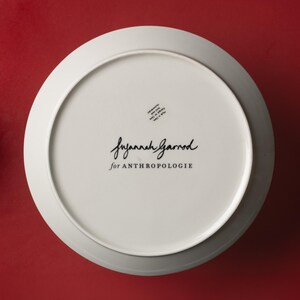 May include: A white ceramic plate with a wide rim, viewed from the underside. The plate features the text "Suzannah Garrod for ANTHROPOLOGIE" in black script. A small sticker is visible near the top. The background is a solid red.