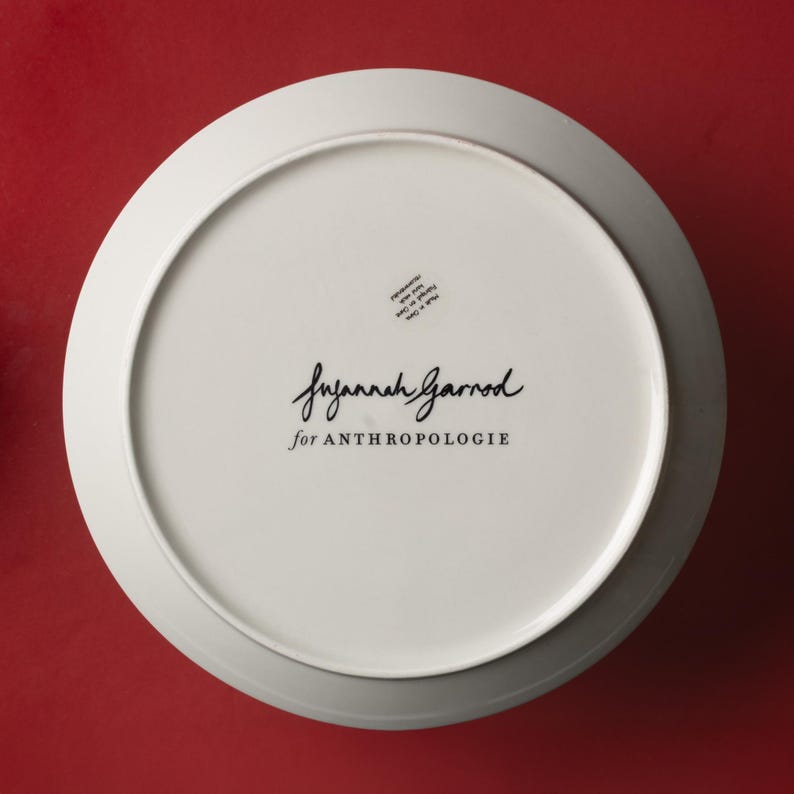 May include: A white ceramic plate with a wide rim, viewed from the underside. The plate features the text "Suzannah Garrod for ANTHROPOLOGIE" in black script. A small sticker is visible near the top. The background is a solid red.