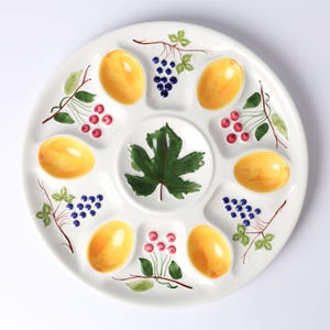 May include: White ceramic serving platter with eight compartments and a central well. The platter is decorated with yellow fruit, green leaves, red berries, and purple grapes. A decorative piece for serving appetizers or snacks.