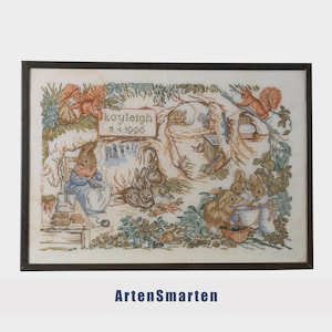 May include: Framed cross-stitch artwork featuring a woodland scene with rabbits and squirrels. The artwork includes the name "Kayleigh" and the date "11.4.1996". The frame is dark brown.