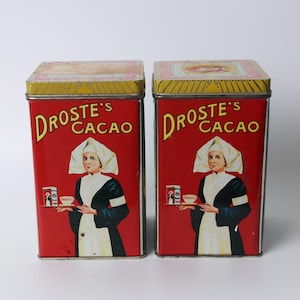 May include: Two vintage Droste's Cacao tins. Each tin is rectangular with a red background and a yellow top. The tins feature a woman in a white bonnet and a black and white dress. The text "Droste's Cacao" is in yellow.