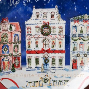 May include: A decorative plate featuring a winter village scene with three detailed houses. The houses are white with red accents, adorned with wreaths, bows, and festive decorations. The background is a dark blue with white stars, and the plate has the text "Up on the".