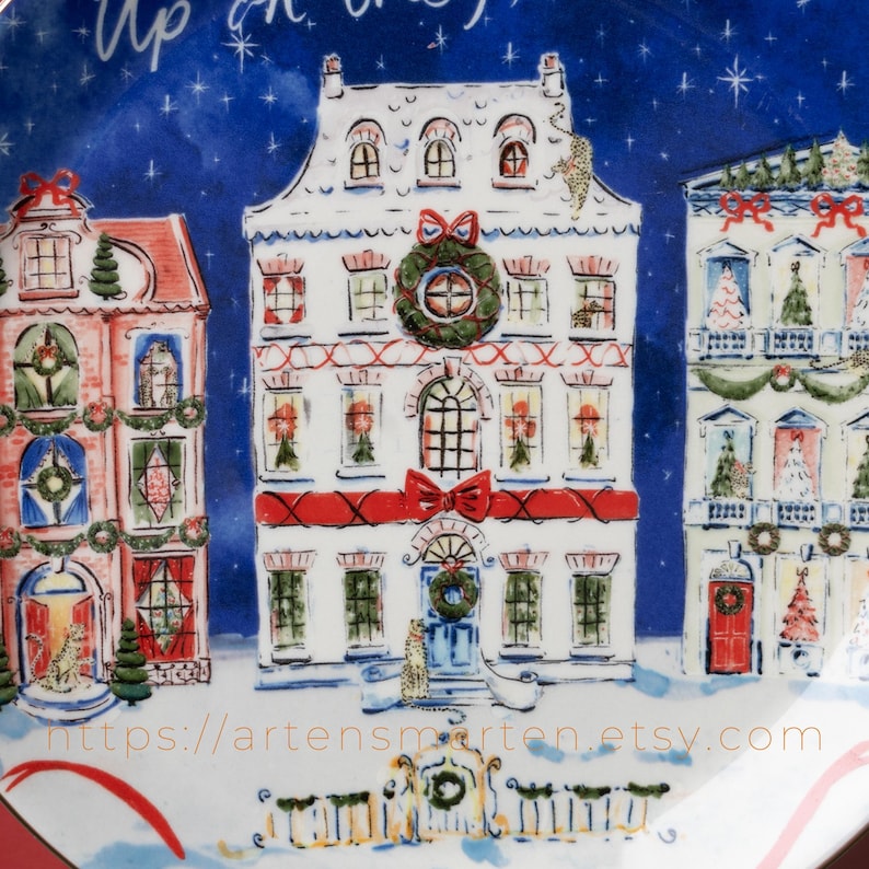 May include: A decorative plate featuring a winter village scene with three detailed houses. The houses are white with red accents, adorned with wreaths, bows, and festive decorations. The background is a dark blue with white stars, and the plate has the text "Up on the".