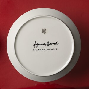 May include: A white ceramic plate with a wide rim, featuring the text "Suzannah Garrod for ANTHROPOLOGIE" in black script. The plate is set against a red background.