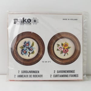 Peut inclure: Un paquet de deux anneaux de rideaux en bois, chacun avec un motif floral brodé. L'un présente des fleurs bleues, l'autre des roses roses et jaunes. Le paquet est étiqueté "pako HANDWERKEN AMSTERDAM - HOLLAND" et "MADE IN HOLLAND". Chaque anneau mesure 7,5 cm de diamètre.