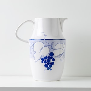 May include: White ceramic pitcher with a curved handle and spout. The pitcher features a blue grape and leaf design around the middle. The design includes a cluster of blue grapes and stylized leaves. The pitcher is suitable for serving beverages.