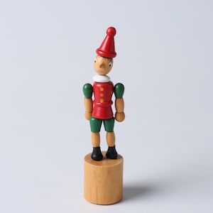 Può includere: Un giocattolo di Pinocchio in legno è in piedi su una base di legno. Il giocattolo è dipinto con un cappello rosso, una giacca e pantaloncini verdi. Le braccia sono verdi e le scarpe sono nere. Il giocattolo ha un colletto bianco e un naso lungo.