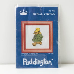 Peut inclure: Un kit de point de croix avec un motif de l'ours Paddington. Le kit comprend une image encadrée de Paddington portant un manteau vert et un chapeau jaune. L'emballage est bleu avec le texte "ROYAL CROWN" et "Paddington". L'image mesure 7 x 7 cm.