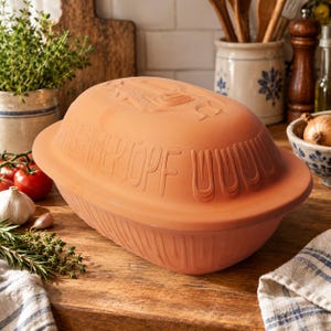 Può includere: Una pirofila ovale in terracotta con coperchio, caratterizzata dalla scritta in rilievo "RÖMERTOPF" sul lato. Il recipiente in terracotta non smaltata color bruno-rossastro presenta un motivo a coste sulla base e un coperchio liscio inciso.