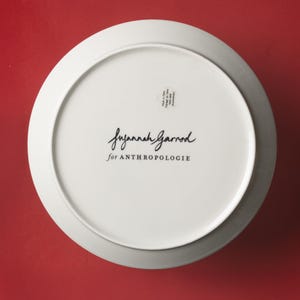 May include: White ceramic plate with a gray rim, viewed from the underside. The plate features the handwritten signature "Suzannah Garrod" and the text "for ANTHROPOLOGIE" in black. A small sticker is visible.