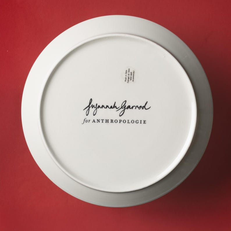 May include: White ceramic plate with a gray rim, viewed from the underside. The plate features the handwritten signature "Suzannah Garrod" and the text "for ANTHROPOLOGIE" in black. A small sticker is visible.