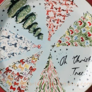 May include: A decorative plate with a light blue background featuring stylized Christmas trees in various colors and patterns. The plate has a gold rim and the words "Oh Christmas Tree" are written in black script.