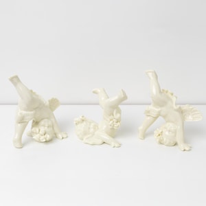 Puede incluir: Tres figuras de querubines de cerámica blanquecina en poses juguetonas. Cada querubín tiene alas y está adornado con detalles florales. Las figuras están dispuestas sobre una superficie blanca, mostrando su diseño caprichoso.