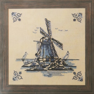 Op de afbeelding: Kruissteekkunstwerk van een blauwe windmolen op een crèmekleurige achtergrond, omlijst in donker hout. De windmolen is gedetailleerd met zeilen en de scène bevat vogels en water. Decoratieve blauwe hoekaccenten.